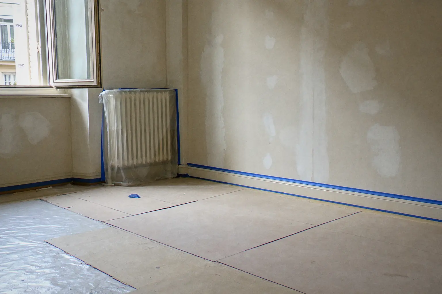 Room protected before painting: parquet covered with sheets and cardboard, wrapped radiator, blue tape along the skirting