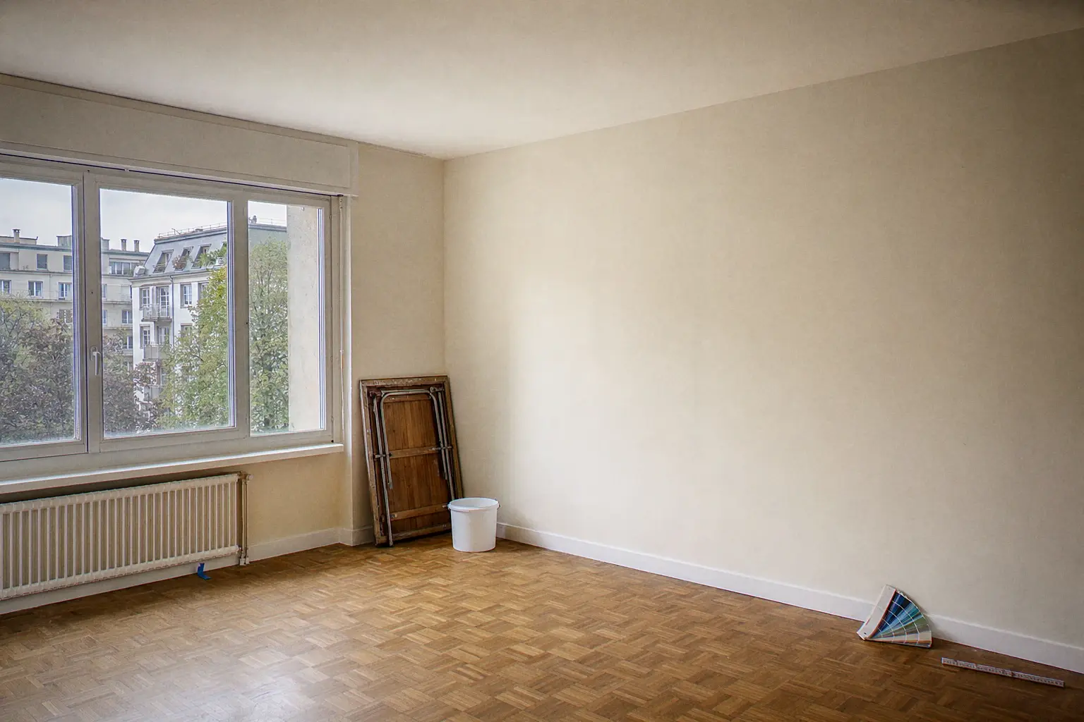 Freshly repainted and handed-over living room in a Geneva apartment: oak parquet, off-white walls, pure white ceiling, colour fan deck and folding rule resting against the wall, folded pasting table at the back