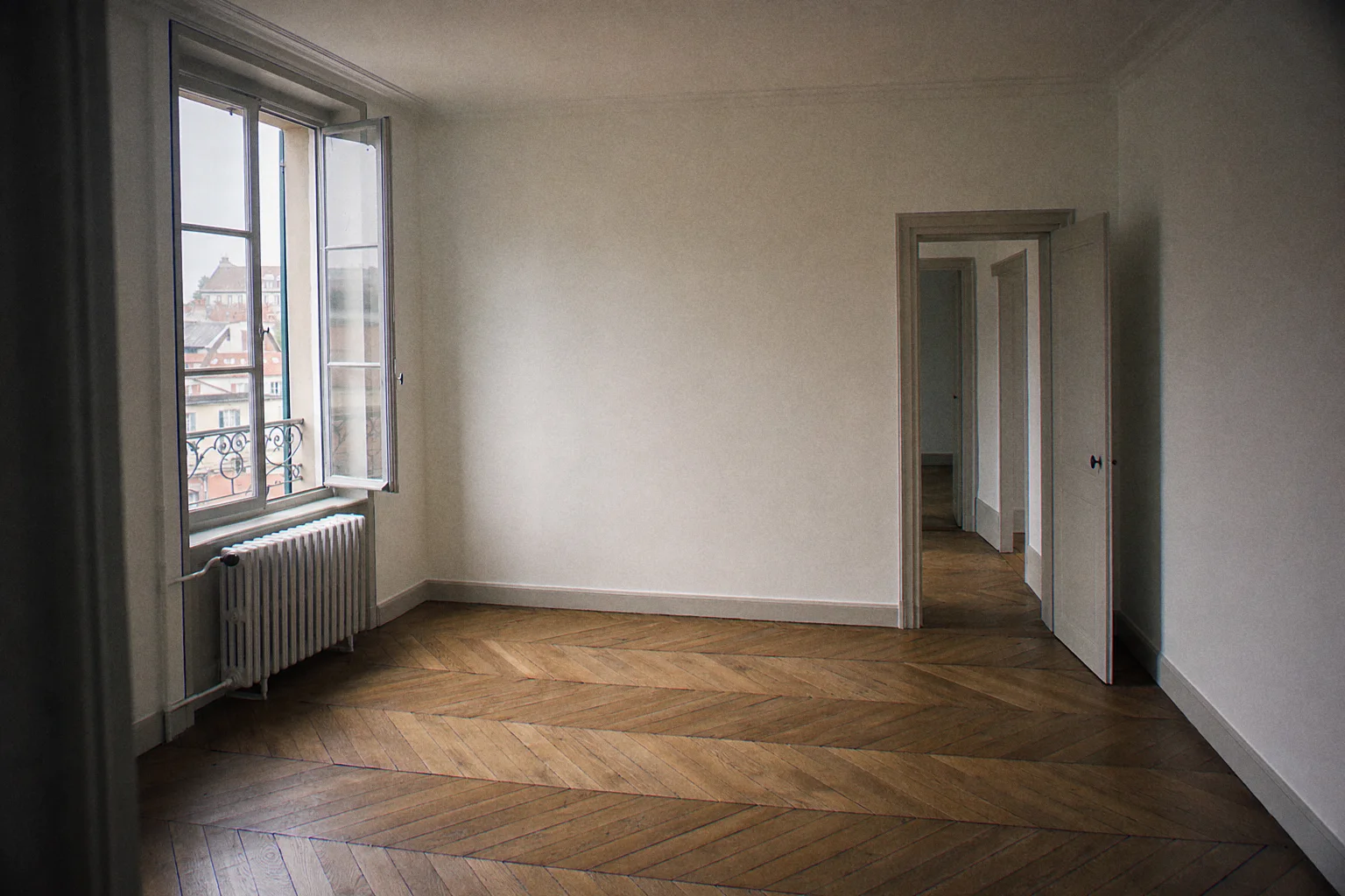Empty living room of an old apartment refreshed between two tenants: freshly repainted off-white walls, repainted cast-iron radiator, herringbone parquet, window ajar for drying